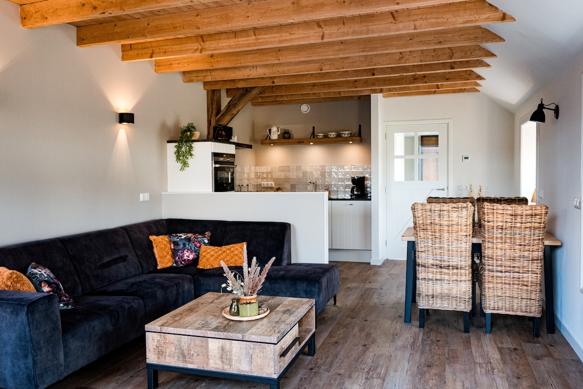 The living area features a sectional sofa in dark fabric, complemented by decorative cushions. A rustic wooden coffee table sits at the center, while a dining table with two wicker chairs is positioned nearby. The kitchen area is accessible and equipped with modern appliances.