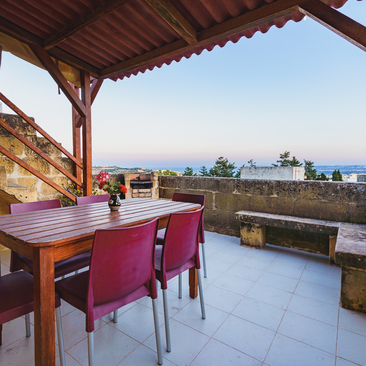 An outdoor dining area is presented, featuring a wooden table surrounded by red chairs. A small flower vase sits at the center of the table. A staircase leads to the upper level, with distant views of the sea and countryside visible in the background.