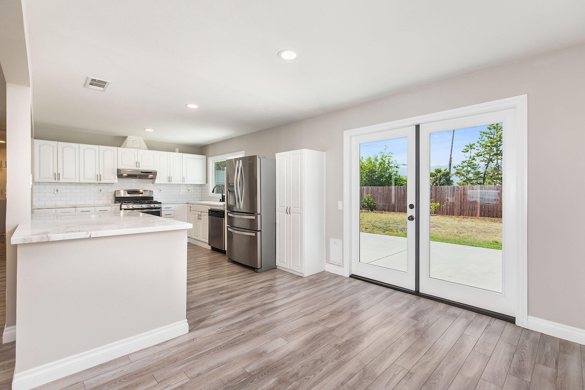 A modern kitchen is visible featuring white cabinetry and stainless steel appliances. Natural light enters through glass double doors leading to an outdoor space. The open layout connects the kitchen to a spacious area with wood-look flooring.