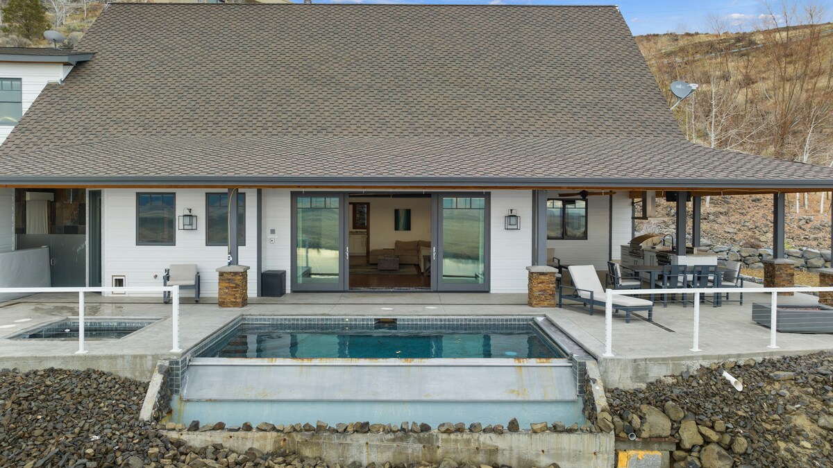 The exterior of the modern beach house is displayed, featuring large glass sliding doors that open to a patio area. An infinity pool is visible at the forefront, surrounded by stone accents, while a spacious dining table and chairs are positioned under the covered area.