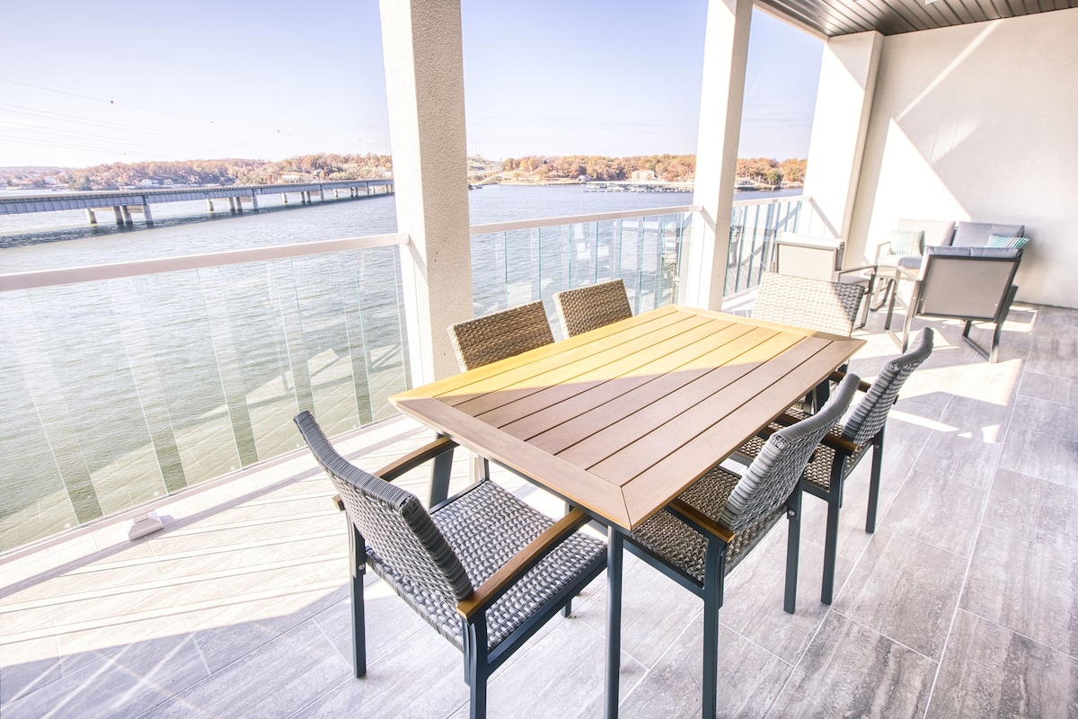 An outdoor deck features a large wooden table surrounded by six woven chairs, overlooking a tranquil body of water. The balcony's glass railing provides panoramic views of the surroundings, enhancing the openness of the space.