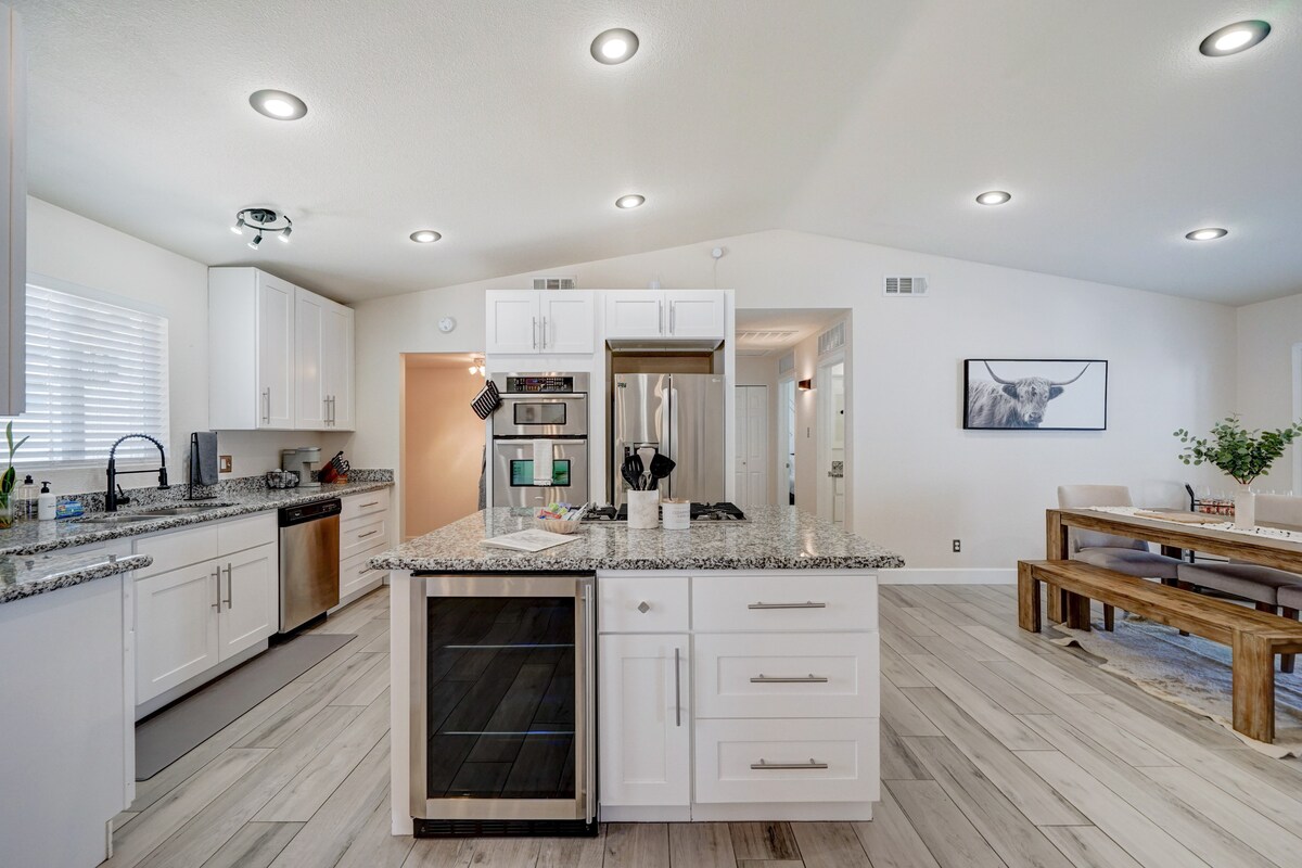 The kitchen features a large central island with a granite countertop, complemented by modern stainless steel appliances. Ample cabinetry offers storage, while natural light filters in through windows. A dining area with a wooden table is visible, creating a cohesive space for meals and gatherings.
