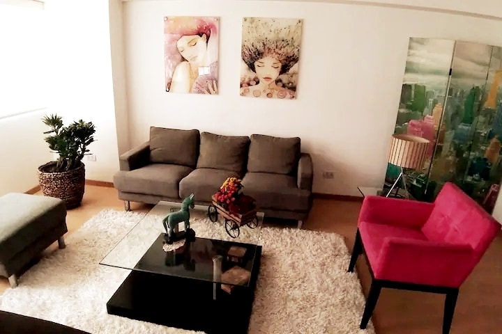 A living area features a large rug, a glass coffee table, and a grey couch complemented by an accent chair in vibrant pink. Two colorful wall art pieces add interest to the space, while a small green plant provides a touch of nature.