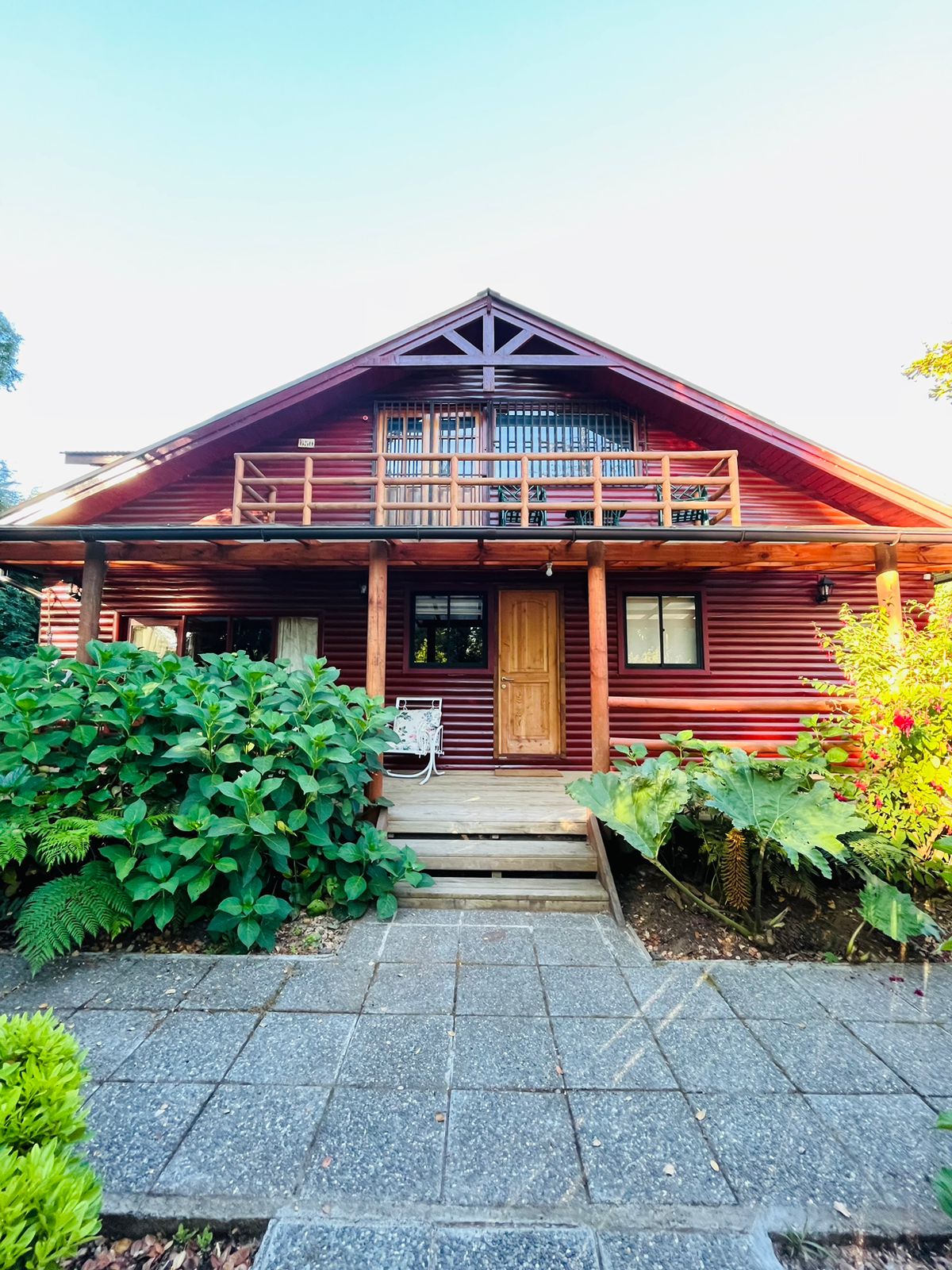 A charming two-story house features a wooden exterior with vibrant red panels. A welcoming entrance is highlighted by a wooden door flanked by windows. The front porch includes a railing and seating area. Lush greenery surrounds the pathway leading to the entrance.
