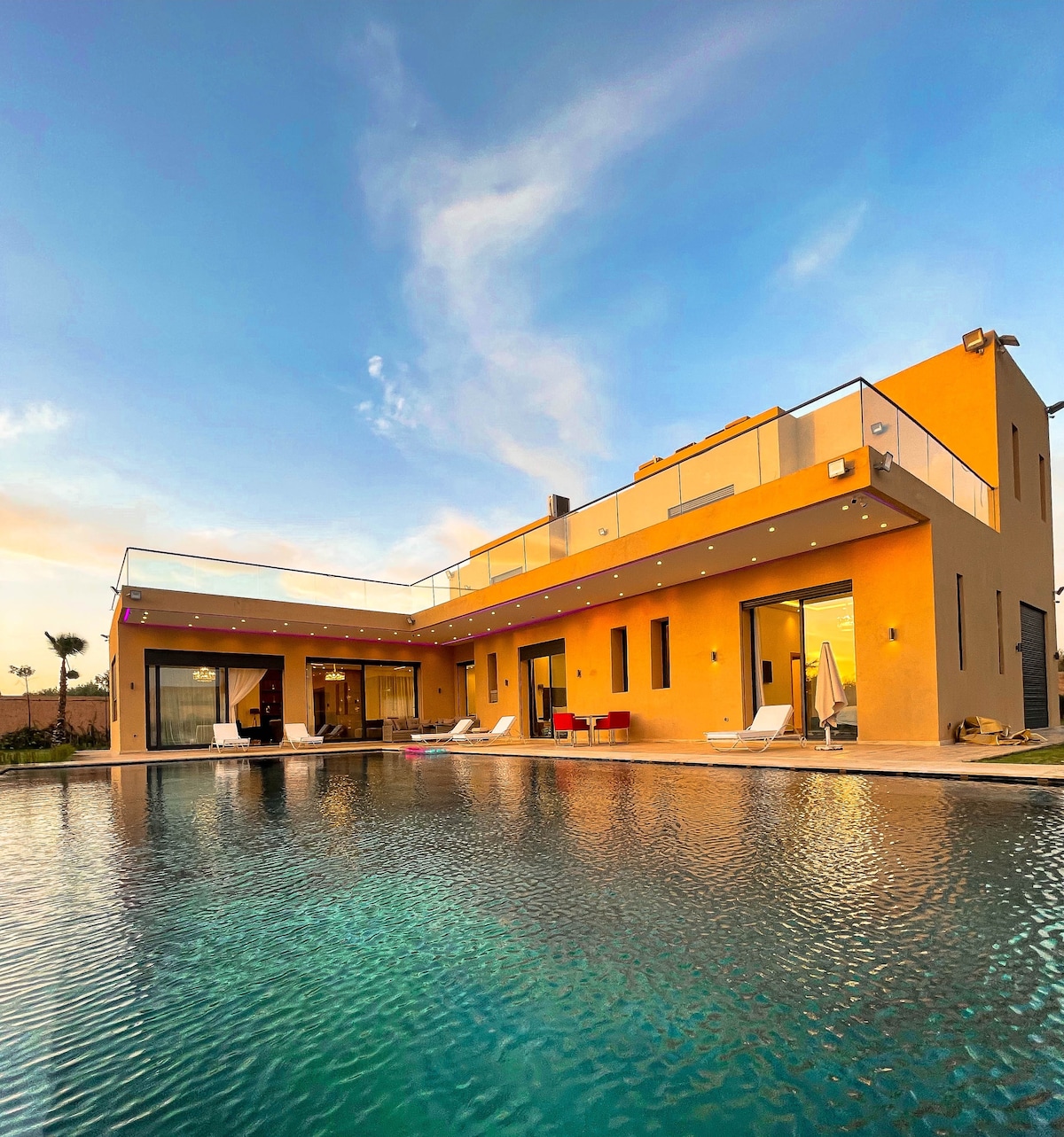 The villa's exterior showcases a modern design with a warm yellow facade. A spacious terrace and large glass doors open to the pool area, reflecting the sky's colors. Lounge chairs are positioned by the inviting pool, which adds to the home's tranquil atmosphere.