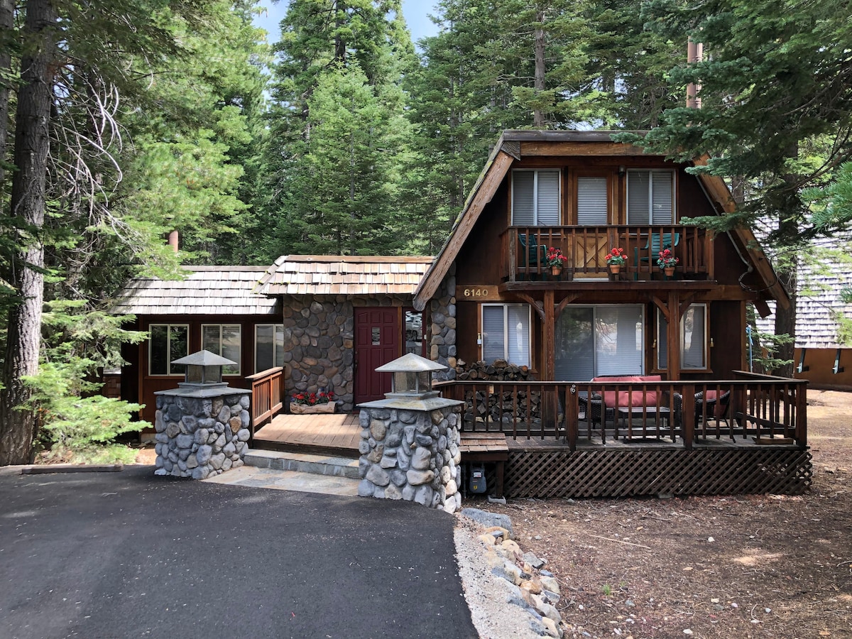 The charming two-story cabin features a stone façade and wooden accents, surrounded by lush trees. A spacious deck with decorative railings and flower boxes adds to the welcome. The entrance is highlighted by a red door, complemented by a well-maintained driveway.