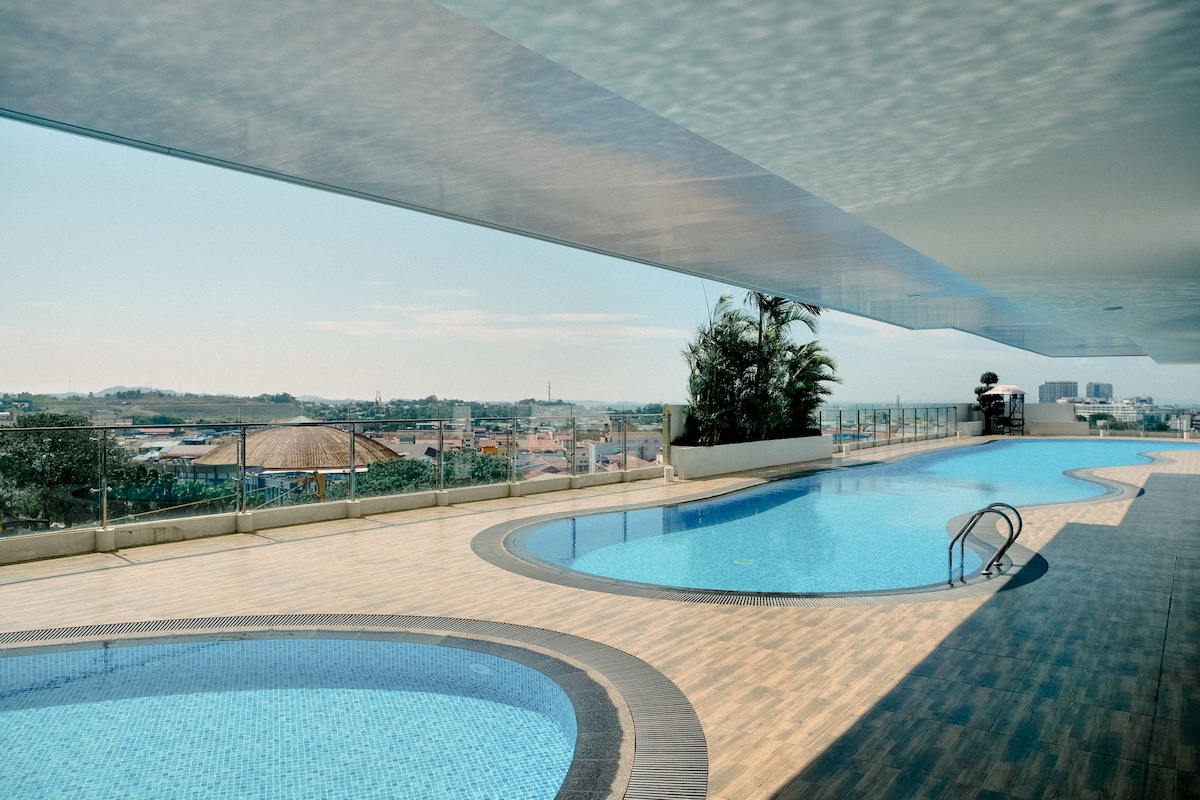 An outdoor pool area with two distinct pools is shown, set against a backdrop of city views. Vibrant sun loungers are placed by the edge, and tropical plants create a serene atmosphere. A sleek overhang provides shade along the poolside, enhancing the open space.
