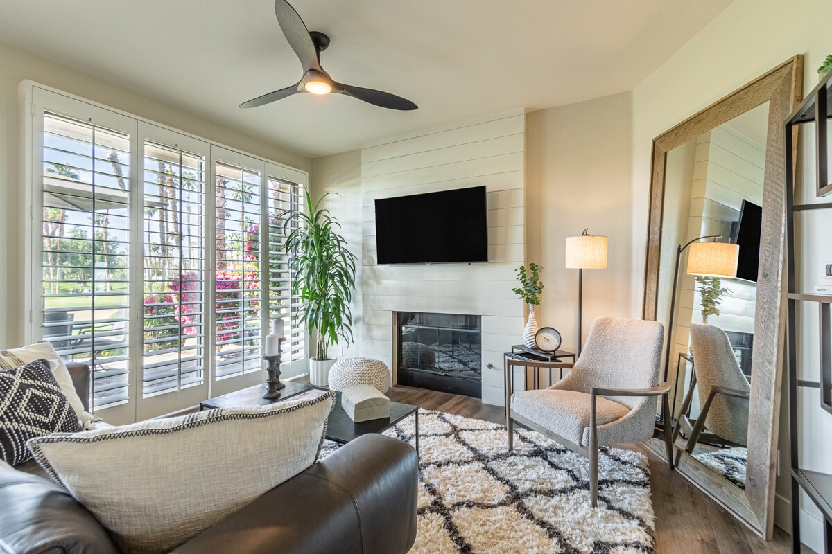 A bright living area features a sleek ceiling fan and large windows with shutters, allowing natural light to enhance the space. A contemporary chair and a plush rug complement the modern decor, while a flat-screen TV is mounted above a stylish shiplap fireplace.