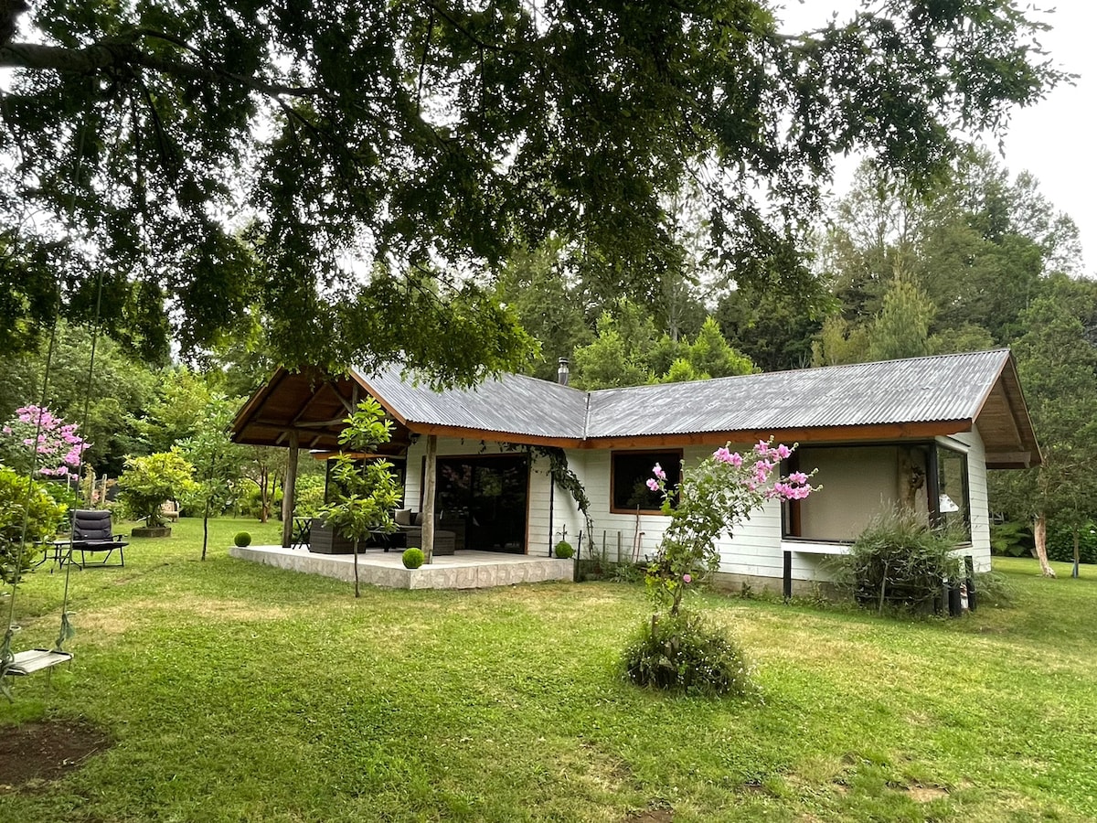 A single-story house with a sloped roof is surrounded by lush greenery and blooming flowers. Outdoor seating is provided on a spacious patio. Large windows open to the garden, offering a seamless connection to the natural surroundings.