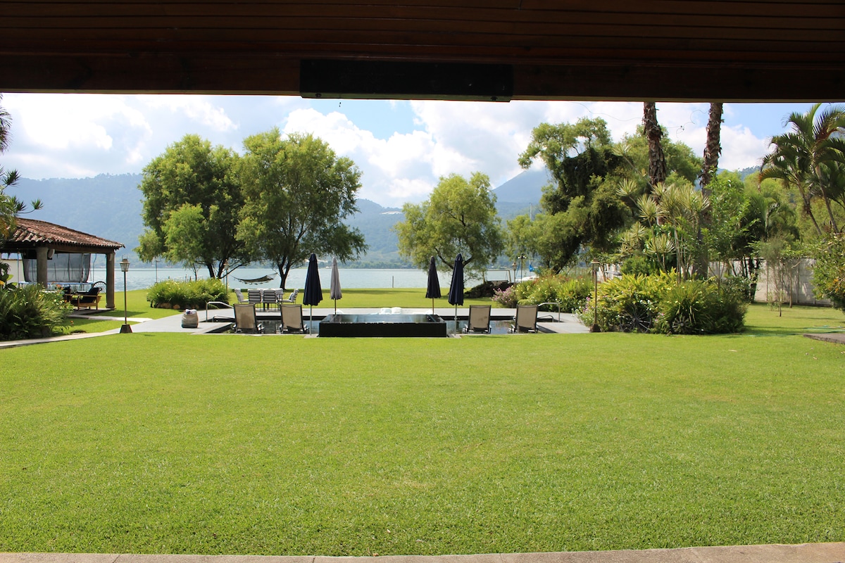 A spacious outdoor area features well-maintained green grass with lounge chairs positioned near a swimming pool. Umbrellas offer shade, while the serene lake and lush trees create a tranquil setting. Mountains can be seen in the background, enhancing the natural landscape.