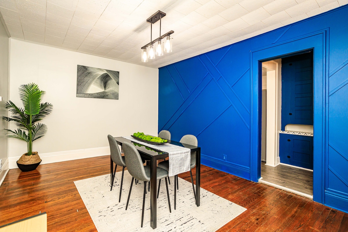 A dining area features a rectangular table set for six, surrounded by modern chairs. A vibrant blue wall serves as a backdrop, contrasting with the warm wooden flooring. A leafy plant stands in the corner, adding a touch of greenery, while a piece of wall art enhances the space.