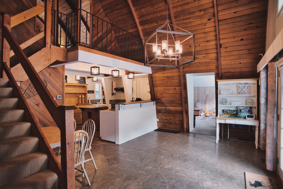 The entryway features a two-level layout with uniquely curved wood walls. A wooden staircase ascends to the upper level, while a simple dining table and chairs are positioned nearby. A modern light fixture illuminates the space, enhancing the cabin's rustic charm.