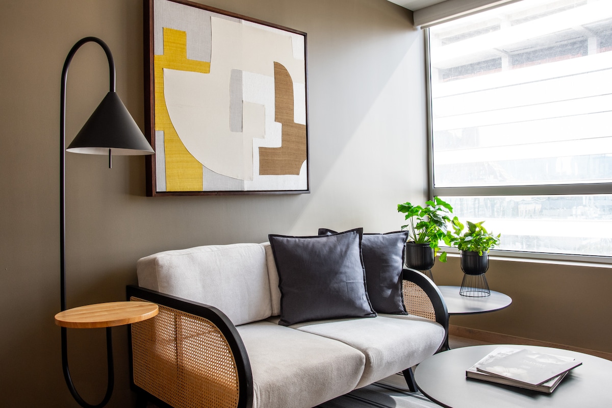 A modern living area features a light-colored sofa adorned with dark cushions, set against a softly painted wall. A geometric artwork hangs above, complemented by a stylish floor lamp. Lush greenery is provided by a potted plant, while round tables complete the inviting atmosphere.