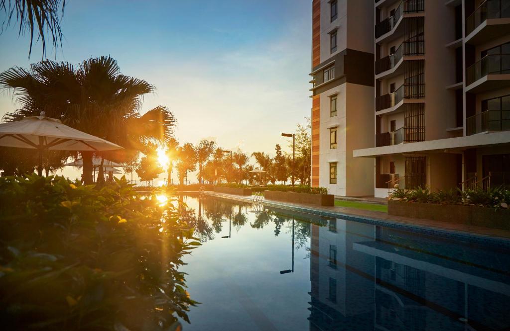A serene swimming pool reflects the warm hues of sunset, surrounded by lush greenery and palm trees. The modern building stands nearby, offering a peaceful setting for relaxation.