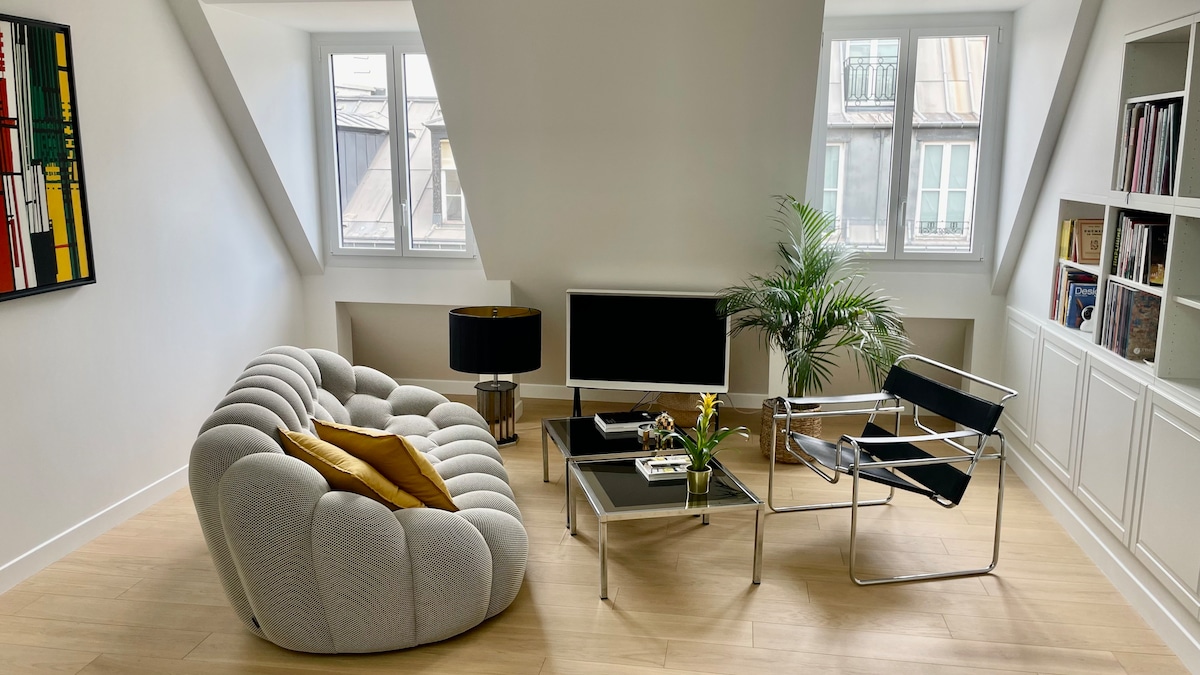 A modern living area features a plush, curved sofa complemented by a bright yellow pillow. A sleek coffee table holds decorative items, while a flat-screen television is positioned on a low stand. Natural light enters through two windows, enhancing the airy ambiance.