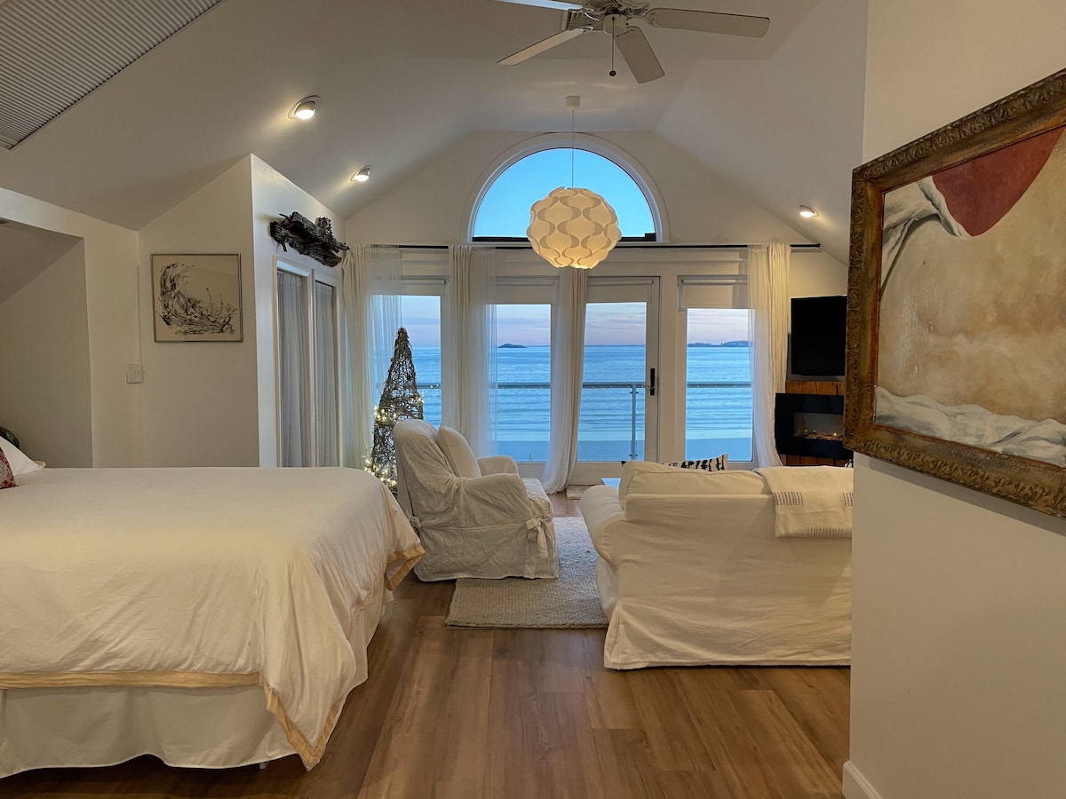 An open loft space features a spacious bed and a cozy seating area. Large windows provide direct views of the ocean, while soft natural light enhances the setting. A decorative light fixture hangs from the ceiling, and a calming palette complements the tranquil atmosphere.