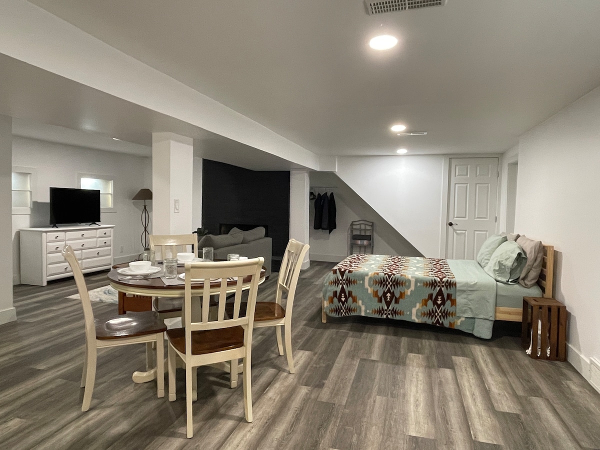 A spacious lower-level area features a queen-sized bed with a patterned blanket and a dining table set for four. A television is visible next to a comfortable seating area with a couch. Light-colored walls enhance the bright, airy ambiance.