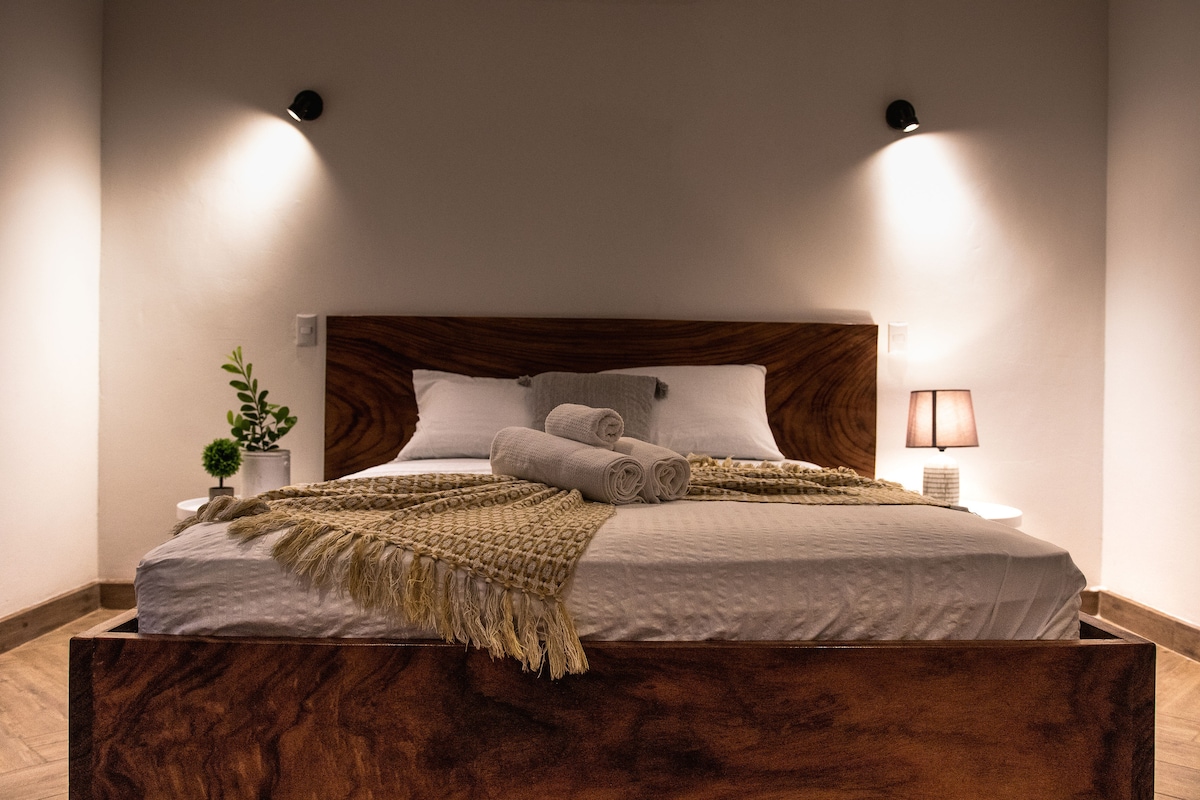 A comfortable bedroom features a large wooden bed with a light-colored blanket draped across the center. Two rolled towels are positioned on top. Soft lighting from wall sconces adds a gentle glow, while a small potted plant and a bedside lamp provide subtle accents.