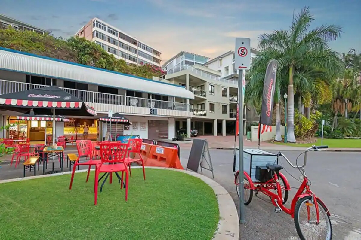An outdoor dining area is visible, featuring rows of red chairs and tables set on grass. A bicycle is parked nearby, alongside a sign indicating a no-parking zone. Various multi-story buildings with balconies form the backdrop, complemented by palm trees.