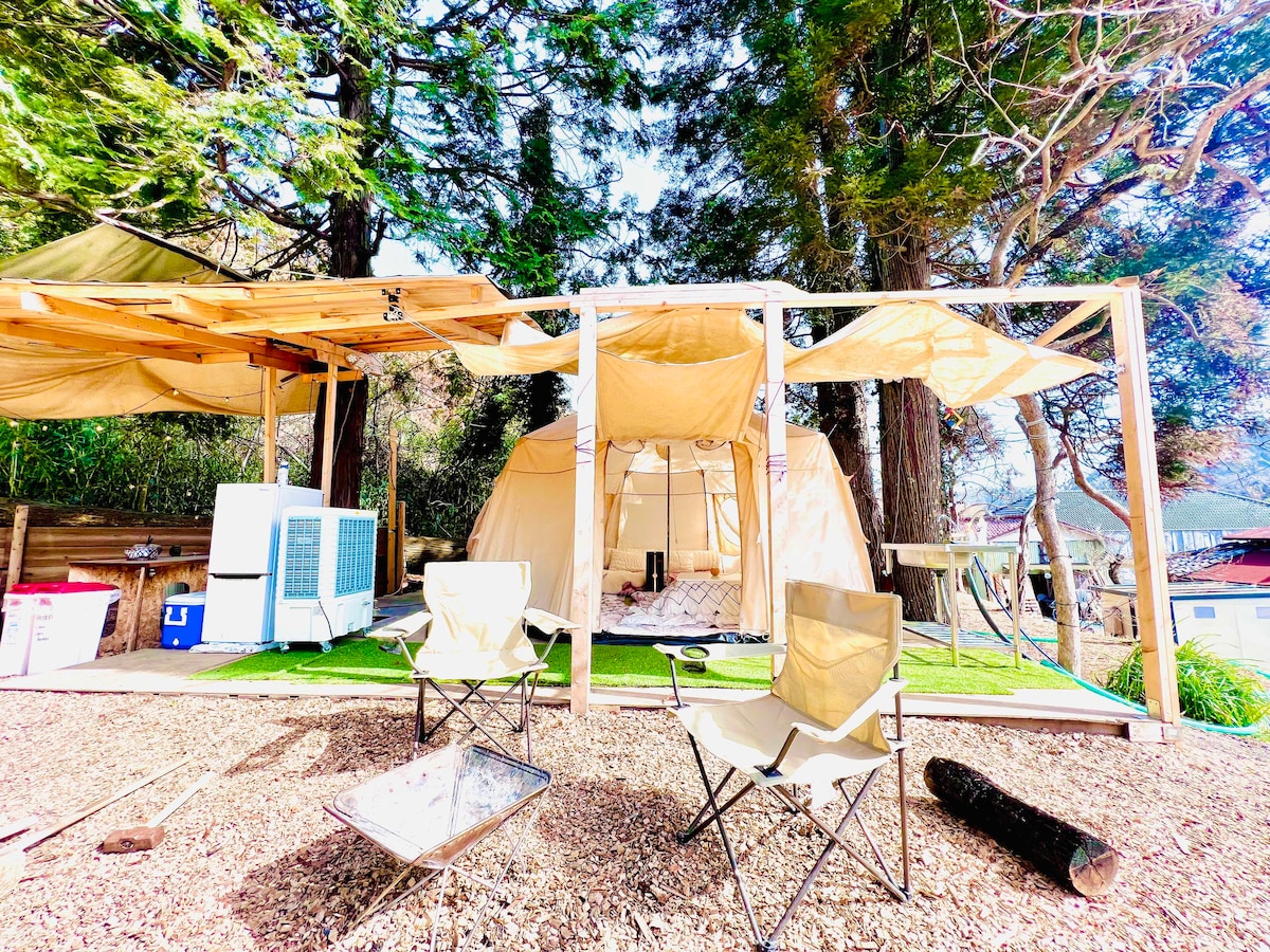 A spacious Nordic tent is set within a natural outdoor environment, surrounded by tall trees. The area features two folding chairs and an outdoor sitting space. A portable air conditioning unit is positioned nearby, contributing to comfort during warmer days.