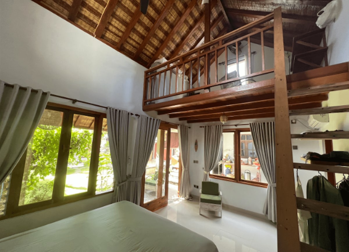 A spacious bedroom features large windows allowing natural light to fill the space. A king-size bed is positioned prominently, with a mezzanine area visible above, accessible via steep stairs. Soft curtains frame the windows, and a small seating area is seen nearby.