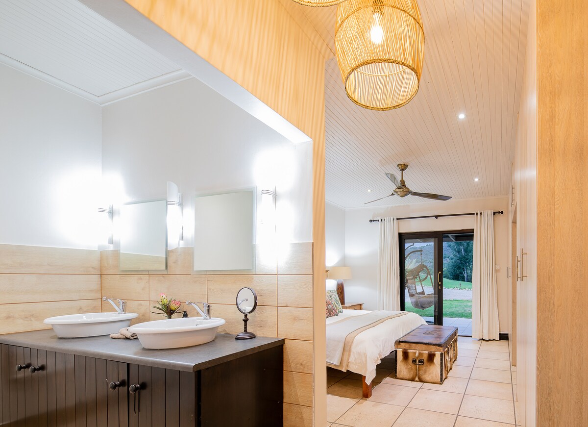 A spacious bedroom with a stylish en-suite area features dual washbasins and mirrors. Natural light filters through sliding glass doors, emphasizing the cozy bed and rustic wooden furniture. Soft pendant lights add a gentle glow to the calming interior.