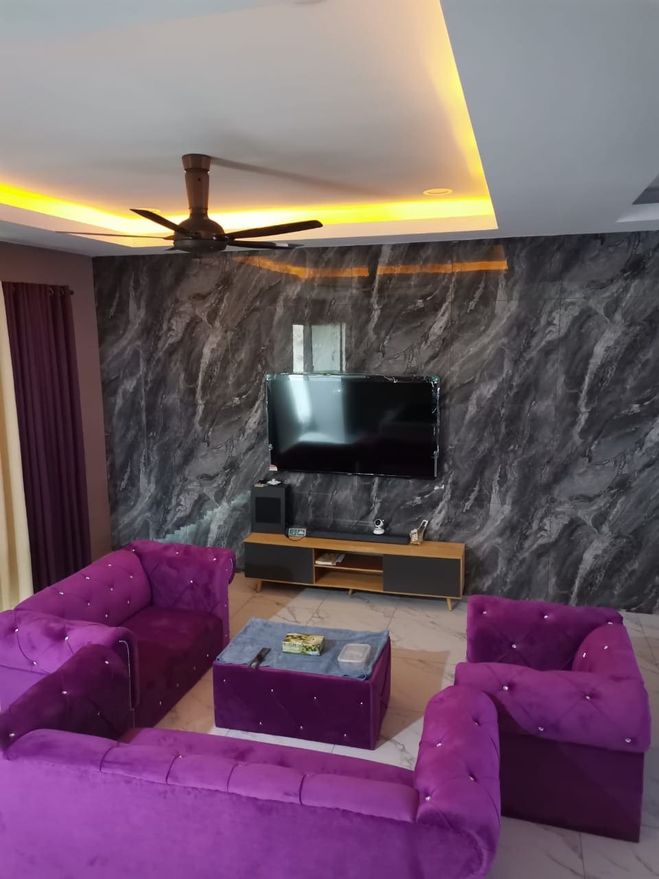 A modern living area features a textured grey wall and a mounted television. Two plush purple chairs and a matching sofa are arranged around a low coffee table. Bright lighting is provided by a ceiling fan, creating a cozy space.