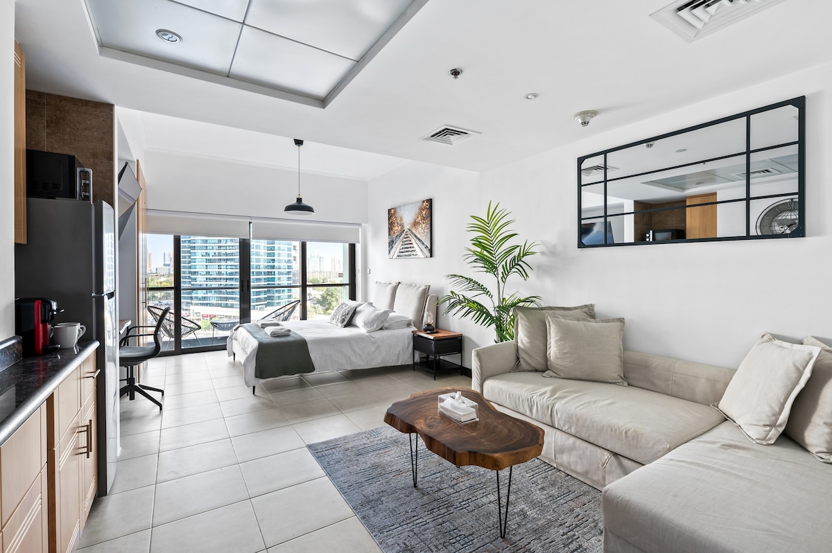 A spacious studio apartment features a comfortable queen-size bed and a cozy sofa that invites relaxation. Natural light floods the space through large windows, offering views of the surrounding area. A stylish coffee table rests on a neutral-toned rug, complementing the modern decor.
