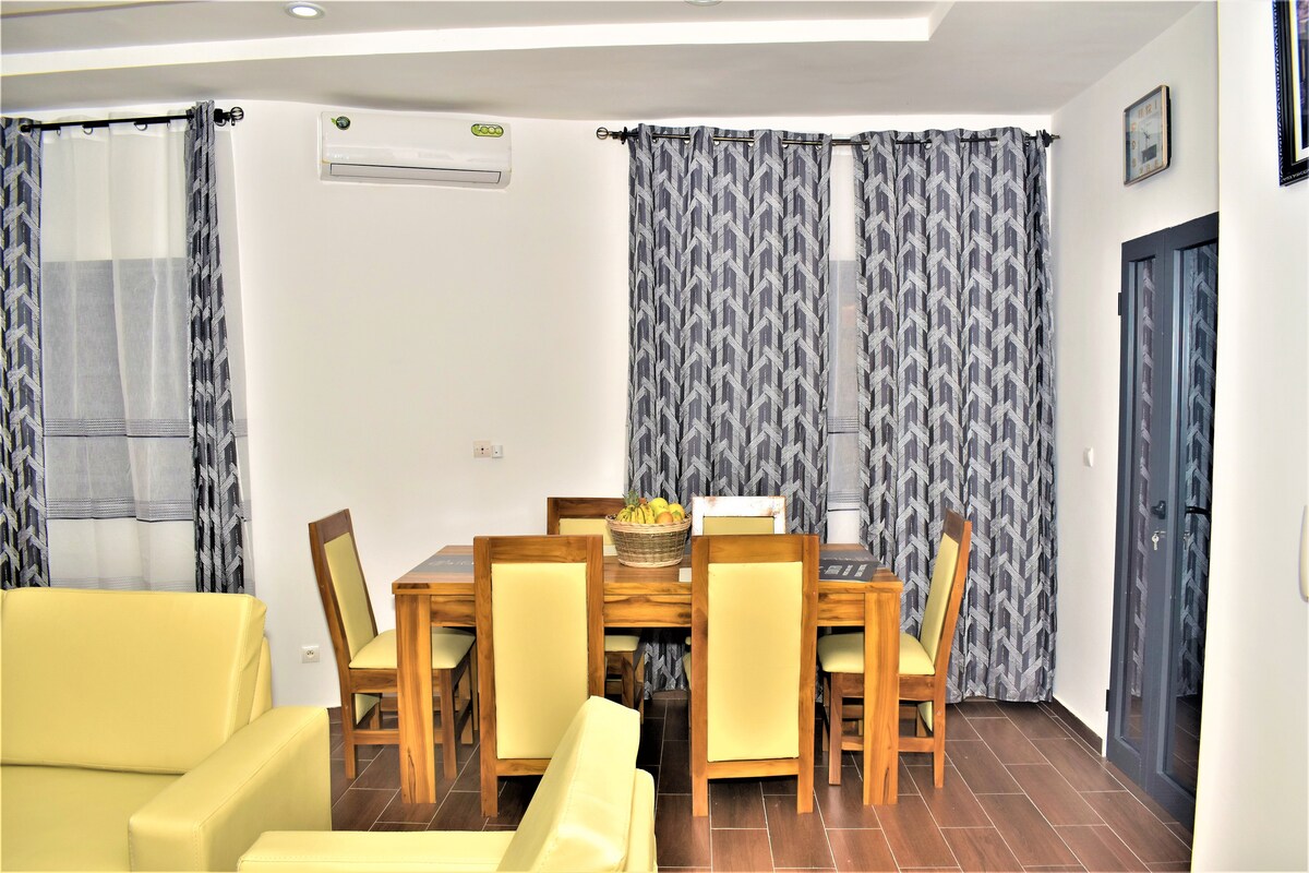 A dining area is presented with a wooden table surrounded by beige chairs. Large curtains in a patterned design frame the windows, and a bowl of fruit is centered on the table. An air conditioning unit is mounted on the wall above.