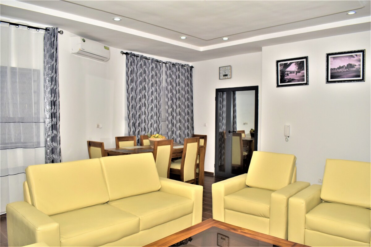 A modern living area is presented, featuring light-colored seating arranged around a glass coffee table. The space is illuminated by recessed ceiling lights, with patterned curtains framing the windows. A dining area is visible in the background, accommodating several chairs and a table.