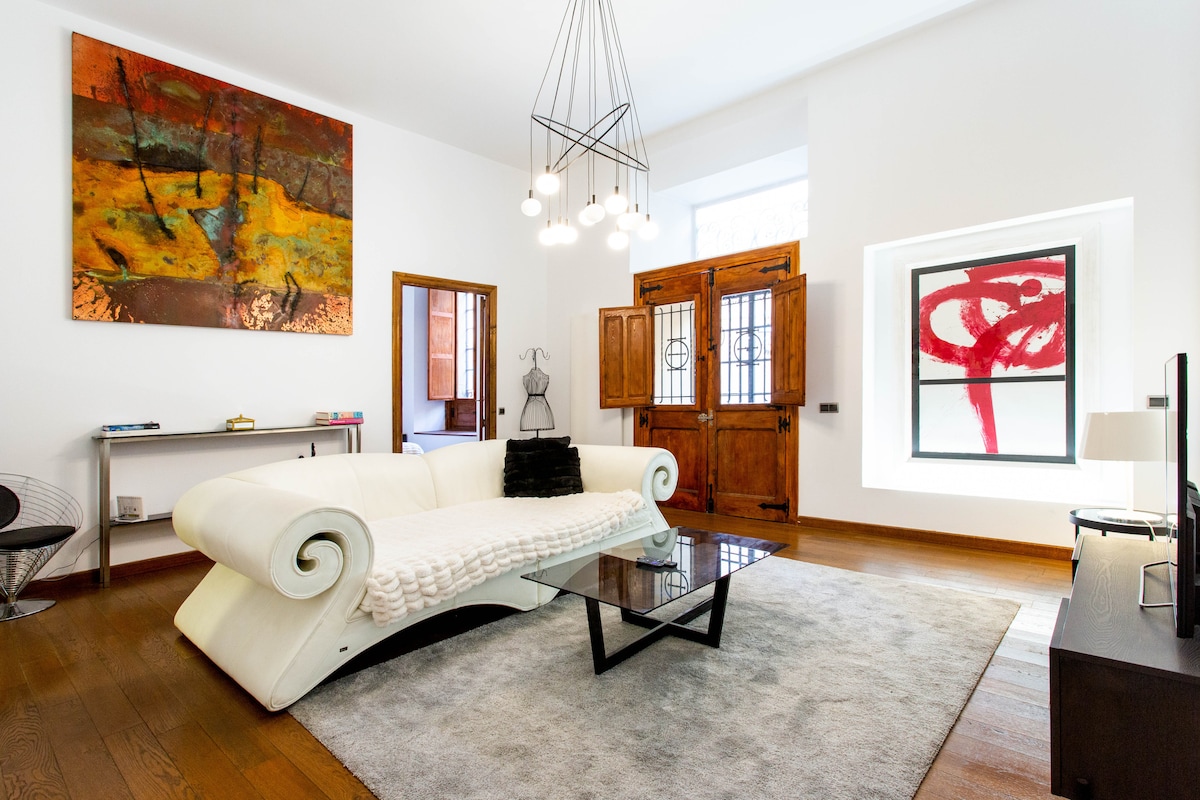 A spacious living area features a unique white curved sofa paired with a modern glass coffee table. A colorful abstract artwork is displayed on the wall, while large wooden doors and a window with decorative glass add character to the room. Soft lighting enhances the ambiance.