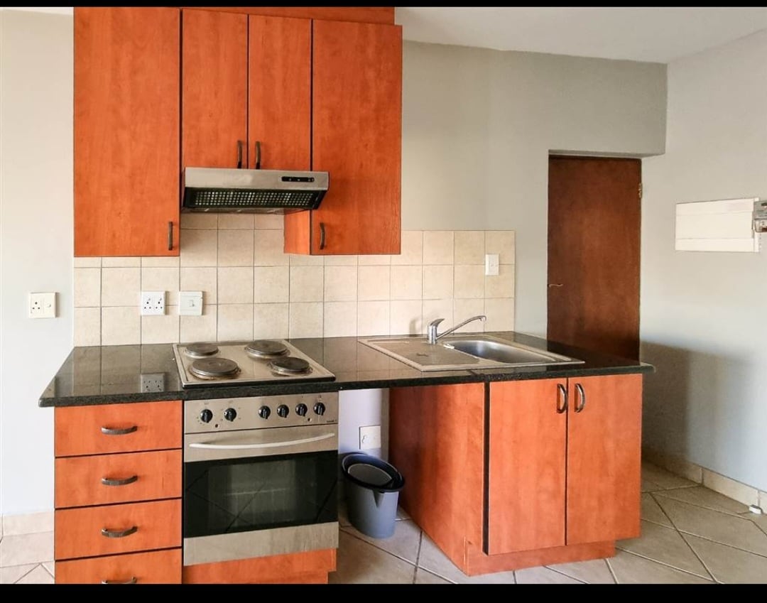 A functional kitchen features wooden cabinetry with a mixture of drawers and cabinets. A stainless steel stove and oven combination is positioned beneath a range hood, while a double-basin sink is located adjacent. The countertop is dark, providing a stylish contrast to the light-colored tiled walls.