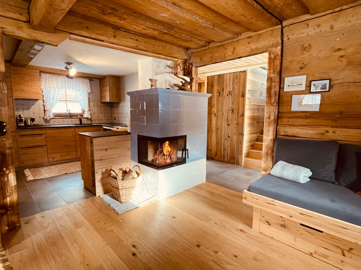 A warm and inviting living area features a modern fireplace set within a rustic stone structure. Light wood paneling complements the space, while a cozy seating area includes a simple couch and a throw pillow. The kitchen area is visible in the background.