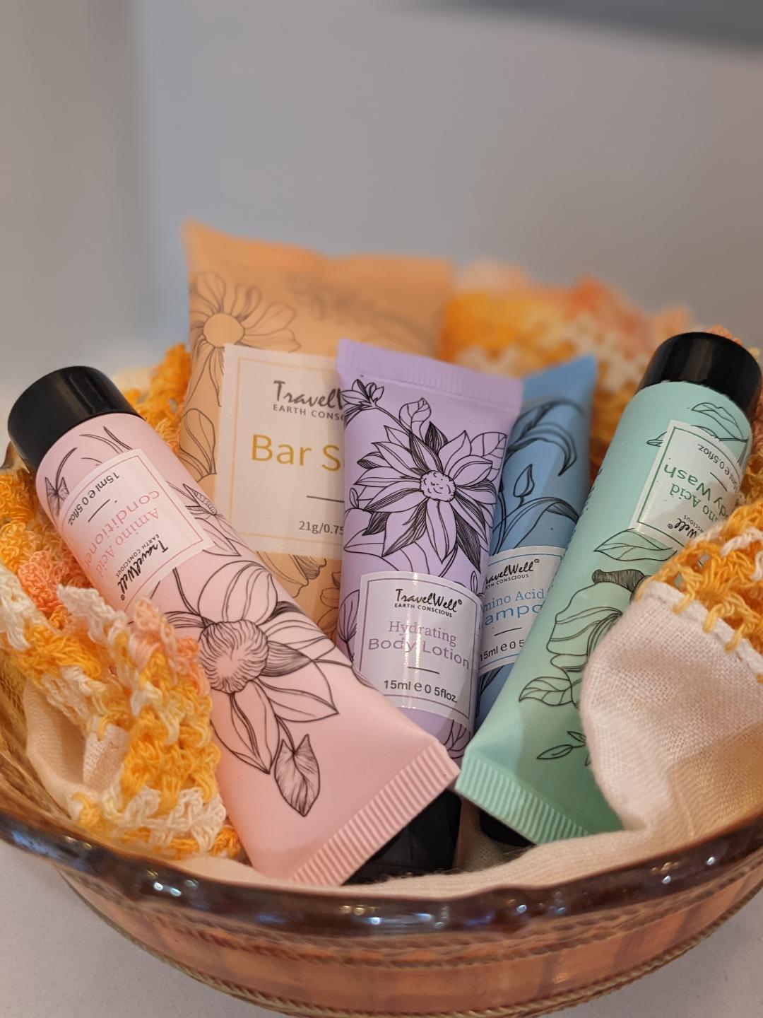 A glass bowl contains an assortment of travel-sized toiletries, including a bar soap, hydrating body lotion, and shampoo. Each item features unique floral designs in pastel colors, arranged amidst a soft fabric liner for added comfort.
