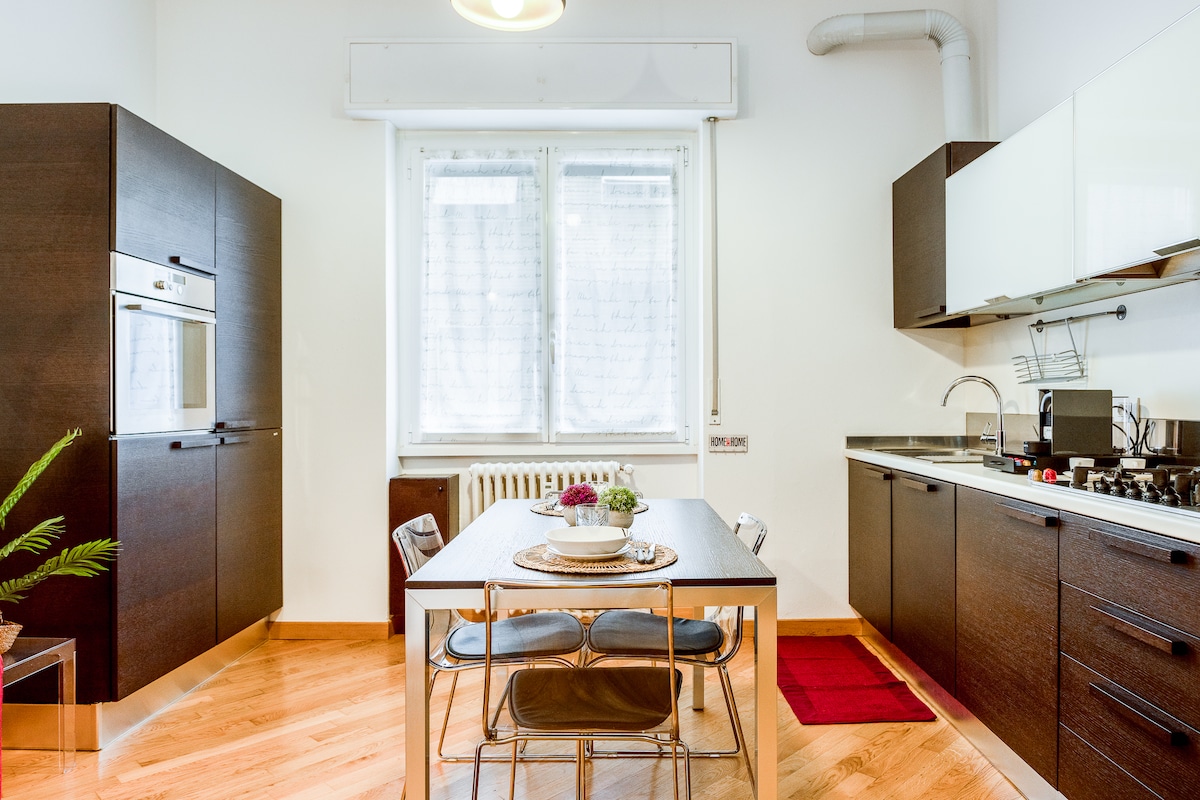 The kitchen features modern cabinetry and a spacious dining table surrounded by transparent chairs. Natural light enters through a window dressed with sheer curtains, illuminating the wood floor. Kitchen appliances and utensils are neatly arranged, providing a functional cooking space.