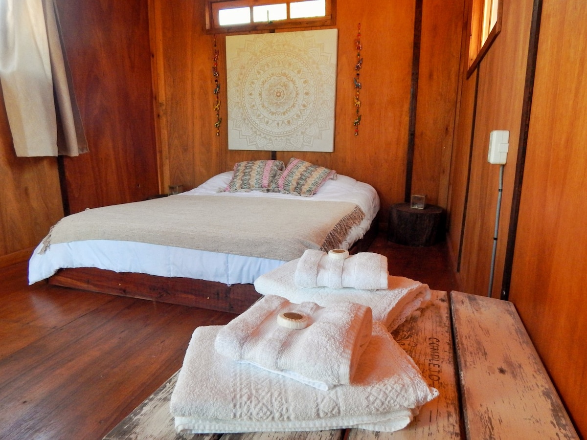 A sleeping area features a spacious bed with decorative pillows, surrounded by wooden walls that create a warm ambiance. Folded towels are neatly arranged on a textured surface, adding a touch of comfort to the setting.