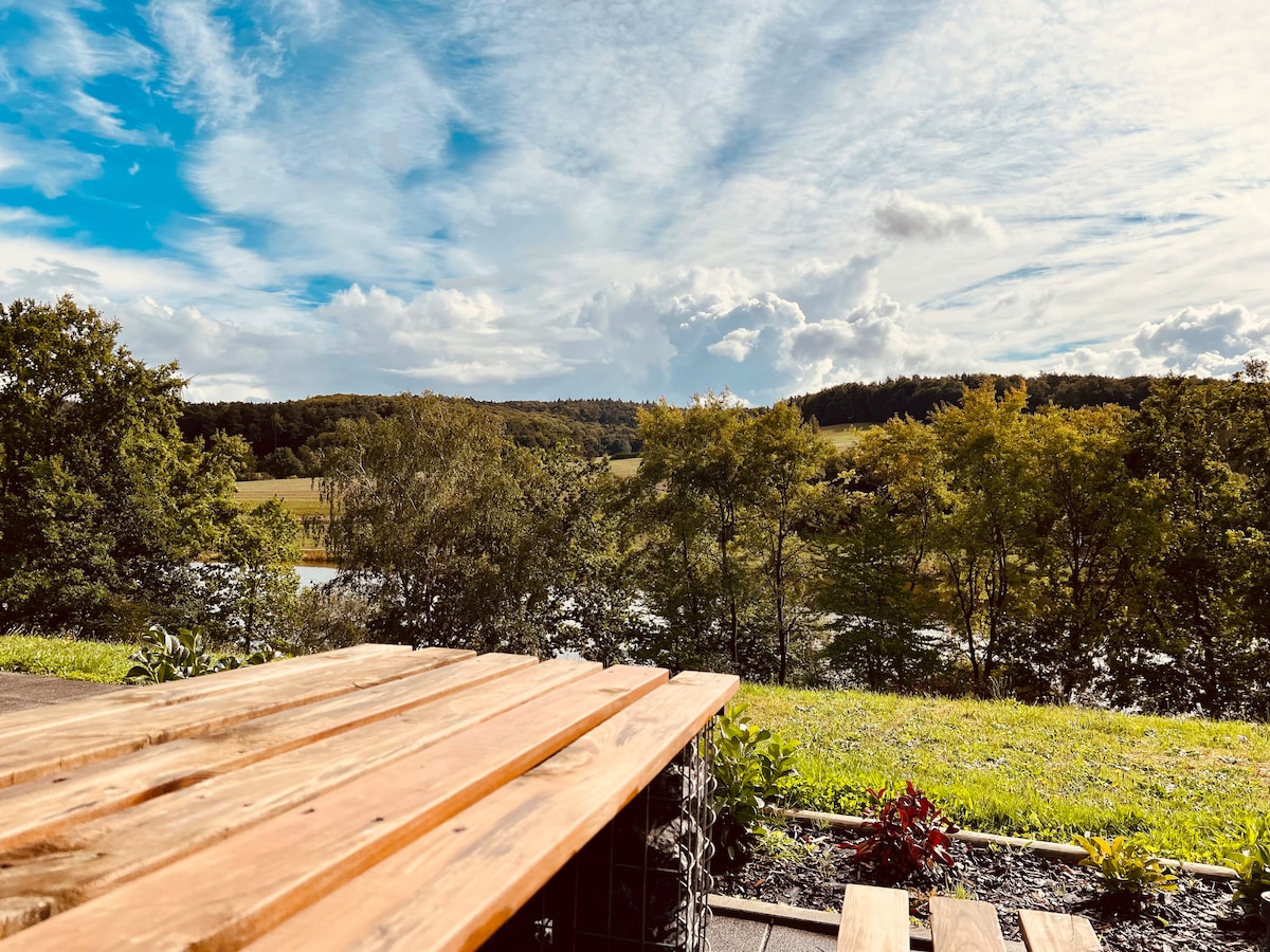 A wooden table is positioned in the foreground, with a view of rolling green hills and a river in the distance. The sky is bright with scattered clouds, and trees frame the landscape, providing a natural ambiance.