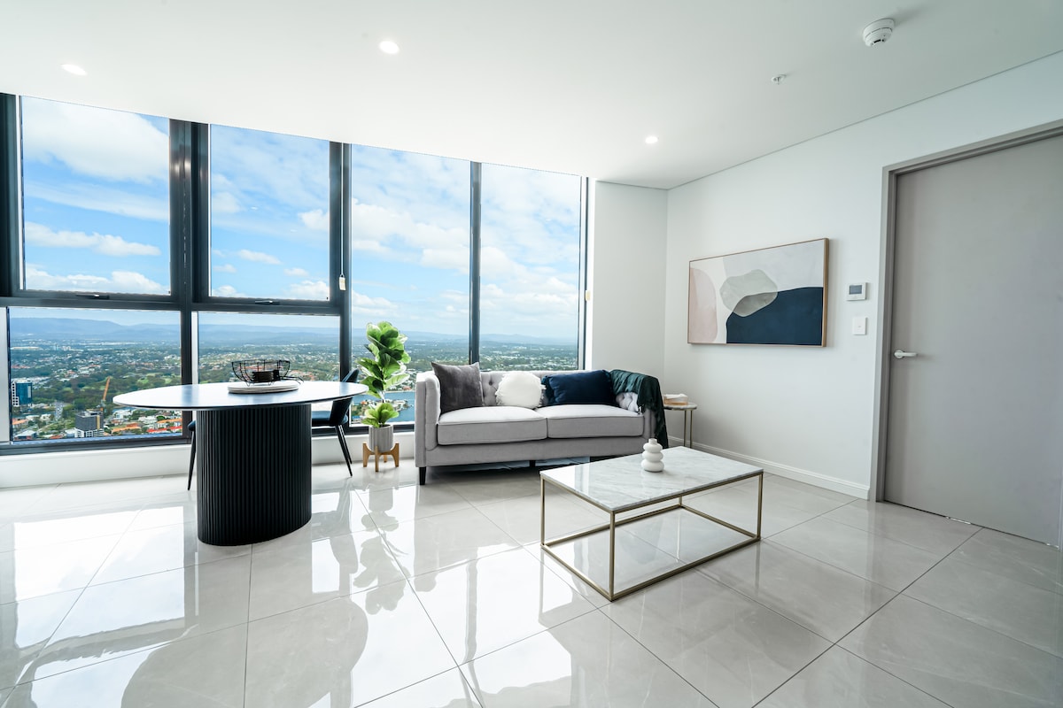 A modern living area showcases large floor-to-ceiling windows, offering expansive views of the city and ocean. A gray sofa is paired with a round dining table and sleek white chairs. A decorative plant and minimalist artwork add subtle touches, while glossy tile flooring reflects natural light.