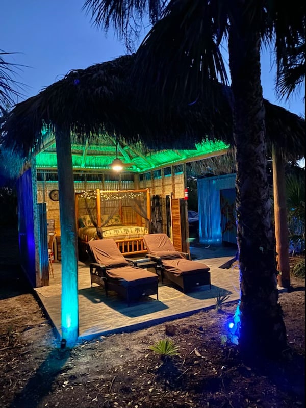 A tropical hut features a thatched roof and ambient lighting, creating a serene atmosphere. Two lounge chairs are positioned under the shelter, and a wooden bed is visible in the background. Palm trees outline the area, enhancing the private outdoor setting.