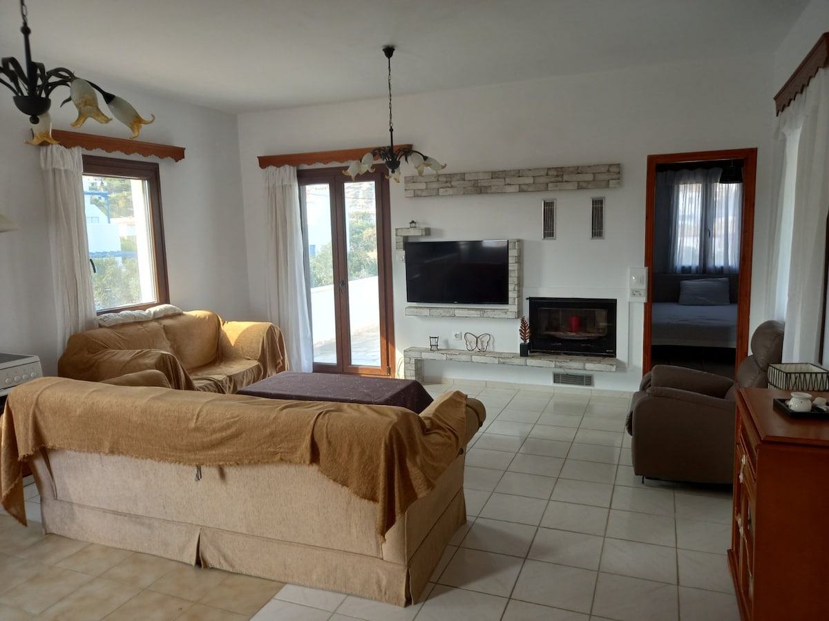 A light-filled living area is furnished with two large sofas draped in neutral covers. A flat-screen television is mounted on the wall opposite, and a modern fireplace offers a cozy focal point. Large windows allow natural light to illuminate the space.