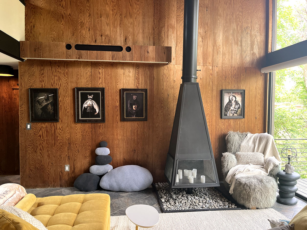 A cozy corner of the living room features a sleek hanging fireplace surrounded by smooth stones and illuminated candles. Stylish art pieces hang on the wood-paneled wall, complemented by plush seating, creating an inviting space for relaxation and conversation.