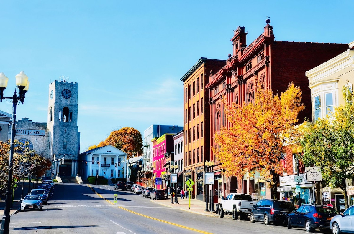 A vibrant streetscape in downtown Geneva showcases historic architecture, featuring a clock tower and a variety of storefronts with colorful facades. Autumn foliage adds warmth to the scene, while parked vehicles are lined along the street. Clear blue skies frame the inviting atmosphere.