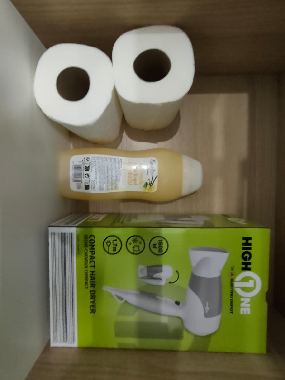 The image displays a neatly arranged shelf featuring two rolls of paper towels, a bottle of shampoo, and a compact hair dryer package. The hair dryer is prominently displayed atop a bright green box, emphasizing its compact size and intended use.