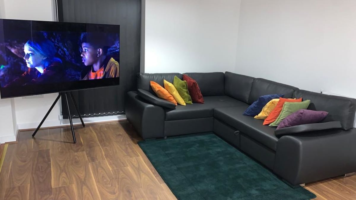 A sectional leather sofa in a neutral color is arranged in the living room, adorned with several colorful cushions. A large flat-screen TV stands on a stand, displaying an image. A plush green rug lies under the seating area, complementing the wooden floor.
