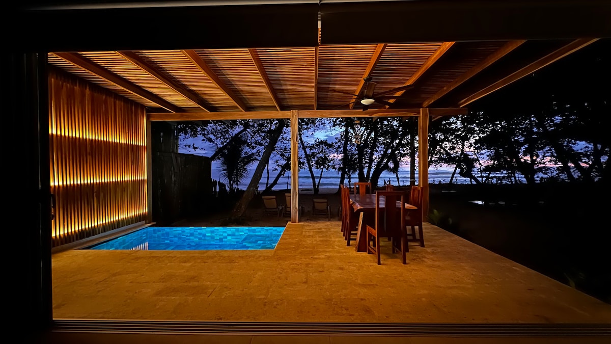 An open-air terrace features a private dipping pool illuminated in soothing blue, complemented by a wooden dining table with chairs. The setting sun casts hues through the trees, creating a serene backdrop of nature just beyond the structure.