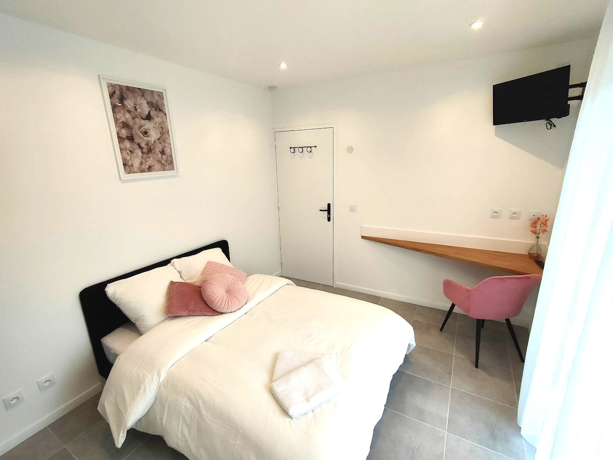 The studio features a cozy sleeping area with a soft bed layered in neutral bedding and decorative pillows. A wall-mounted television is visible above a minimalist desk accompanied by a pink chair. Natural light enters through sheer curtains, illuminating the serene interior.