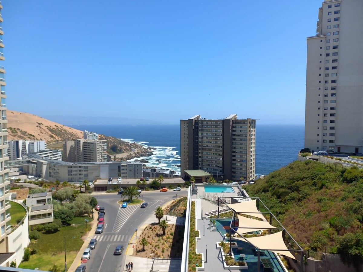 A panoramic view from a high vantage point captures the coastline, featuring the vibrant blue ocean against a backdrop of sandy dunes. Modern buildings line the shore, while landscaped areas and a pool are visible below, offering a glimpse of the communal spaces in the area.