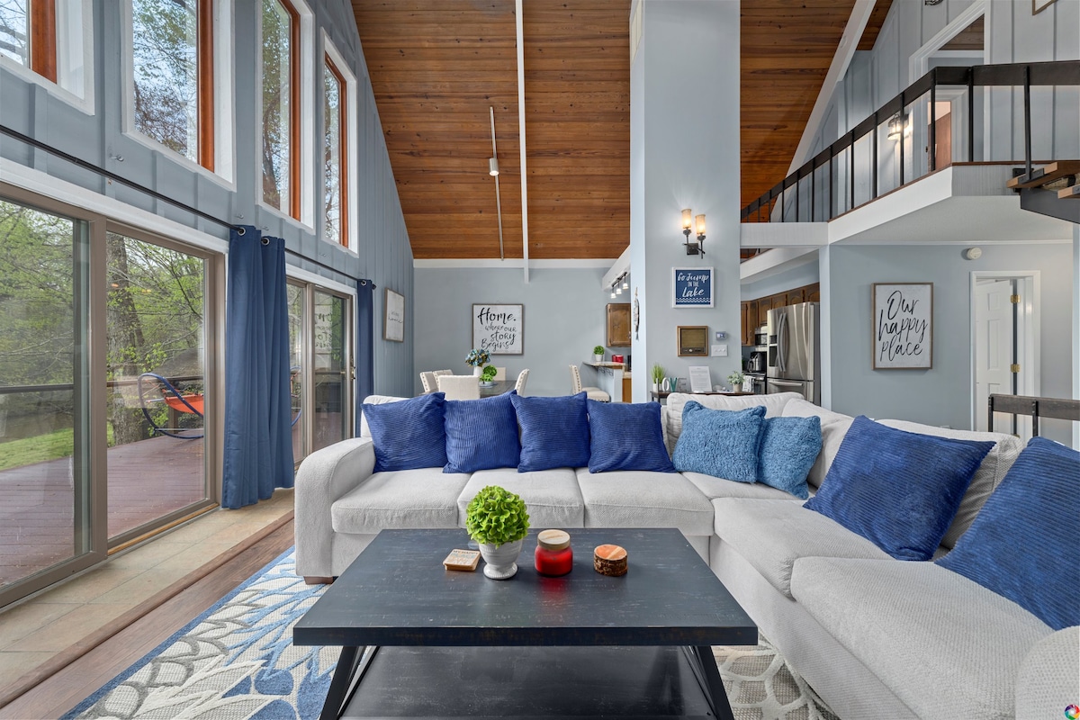 A spacious living area features a large sectional sofa adorned with blue accent pillows, complemented by a black coffee table holding decorative items. High ceilings with exposed beams enhance the open feel, while large windows allow natural light to illuminate the space.