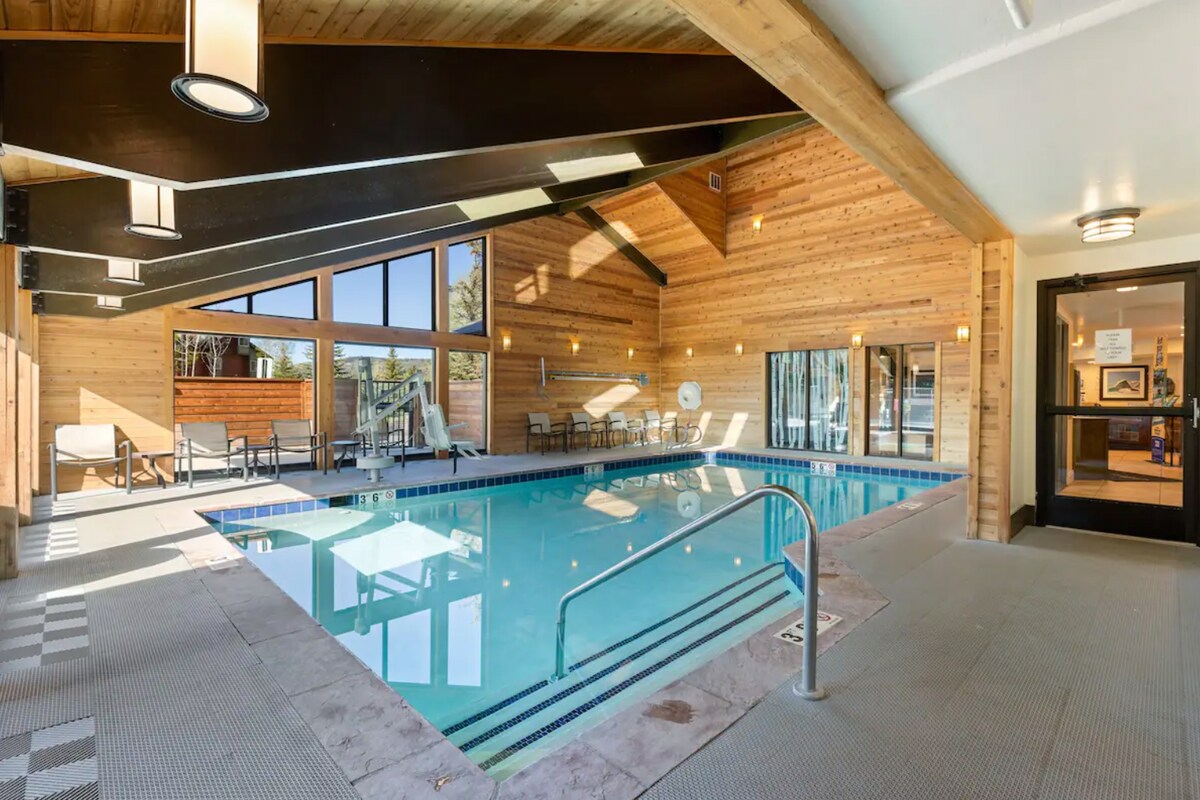 An indoor swimming pool is featured, surrounded by large windows allowing natural light. Wood paneling accents the walls and ceiling, creating a warm atmosphere. Lounge chairs are positioned around the pool, and an accessible entry ramp leads into the water.