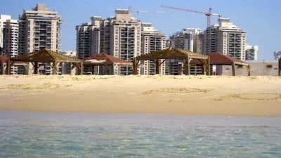 The image captures a sandy beach with several wooden cabanas positioned on the shore. In the background, modern buildings line the horizon, with cranes visible, indicating ongoing development. The calm water reflects the clear blue sky, creating a serene coastal atmosphere.
